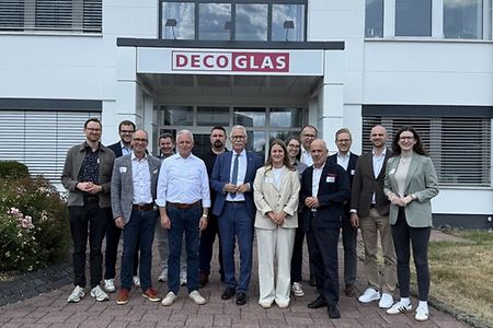 Eine Personengruppe steht vor einem Geb&auml;ude mit dem Schild "DECO GLAS"