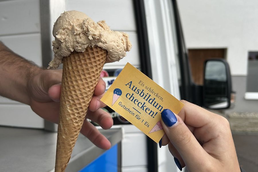 Ein Eisverkäufer reicht eine Kugel Eis in der Waffel an eine Person. Die Person reicht einen Gutschein.