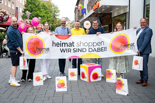 Mehrere Menschen stehen mit einem Banner, Luftballons und Einkaufst&uuml;ten in einer Innenstadt. 