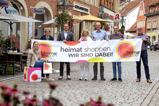 Mehrere Menschen stehen mit einem Banner, Luftballons und Einkaufst&uuml;ten in einer Innenstadt.