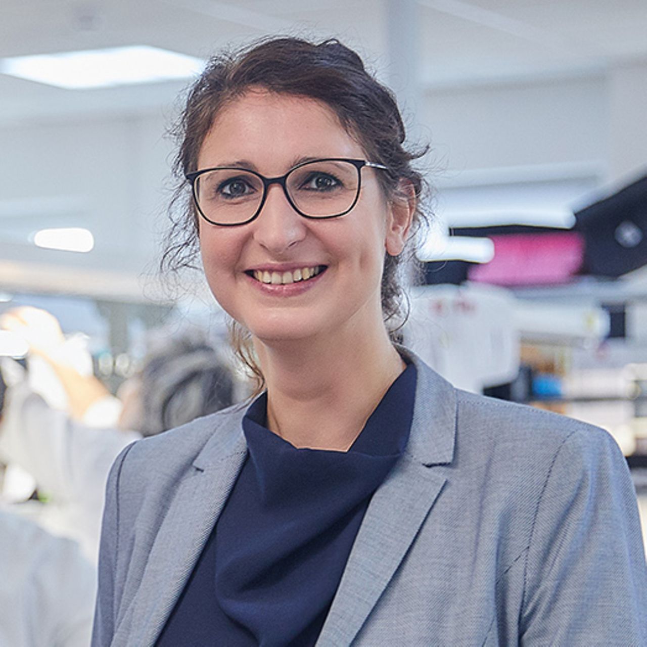Porträt einer Person mit Brille in hellgrauem Blazer und dunklem Oberteil in einer hellen Innenraumumgebung.