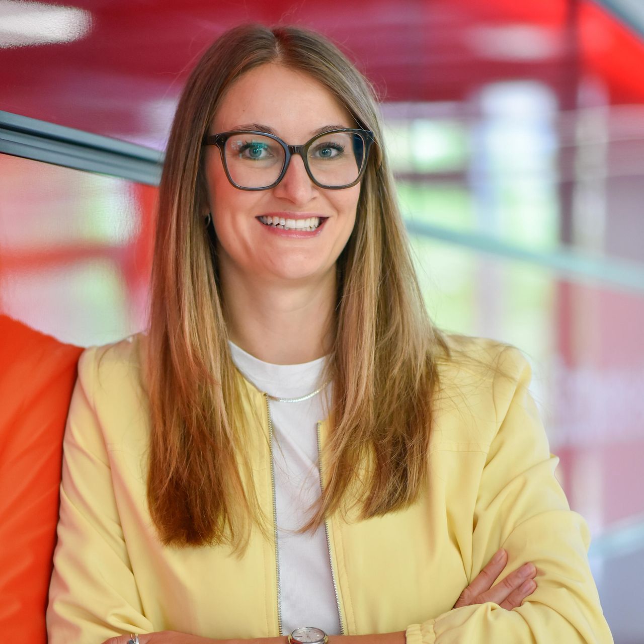 Porträt einer Person mit Brille und hellem Blazer vor einer modernen Innenraumkulisse mit roten Gestaltungselementen.