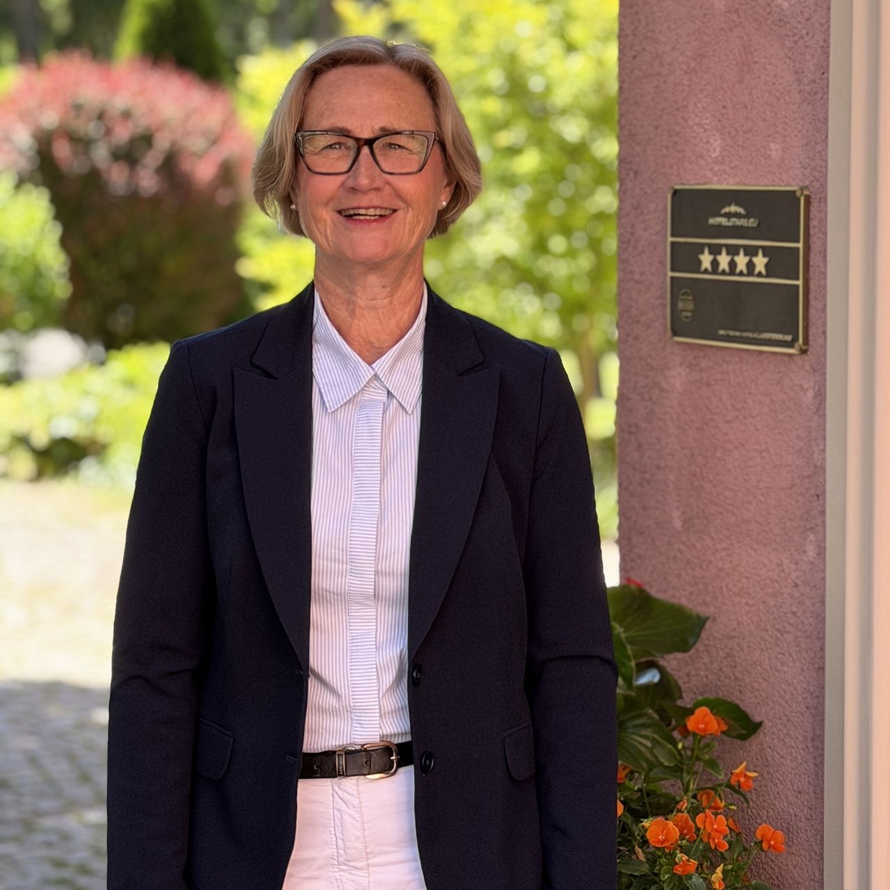 Porträt einer Person mit Brille und dunklem Blazer vor einem Gebäude mit Gartenanlage im Hintergrund.