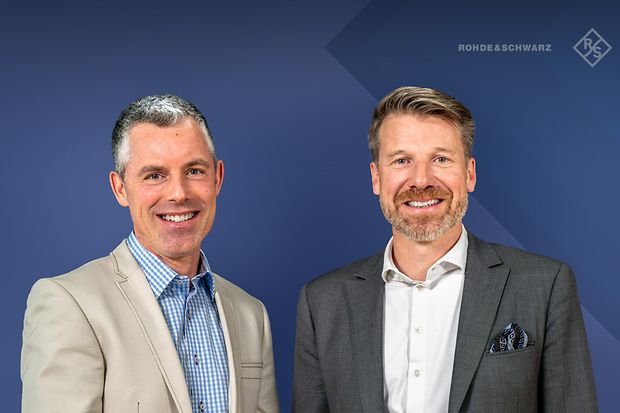 Zwei M&auml;nner im Jackett stehen nebeneinander und l&auml;cheln.