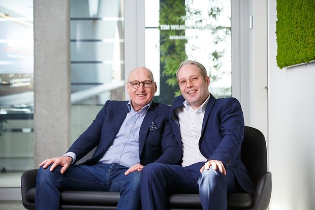 : Zwei M&auml;nner mit Brille, gekleidet in Hemd und Jackett, sitzen auf einem Sofa und l&auml;cheln.