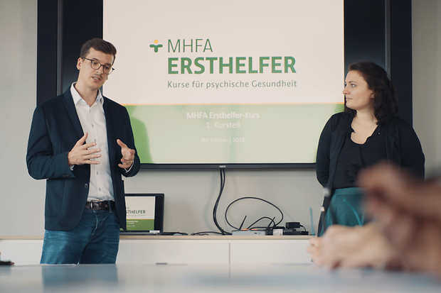 Zwei Menschen stehen vor einer Leinwand mit der Aufschrift "MHFA Ersthelfer" 