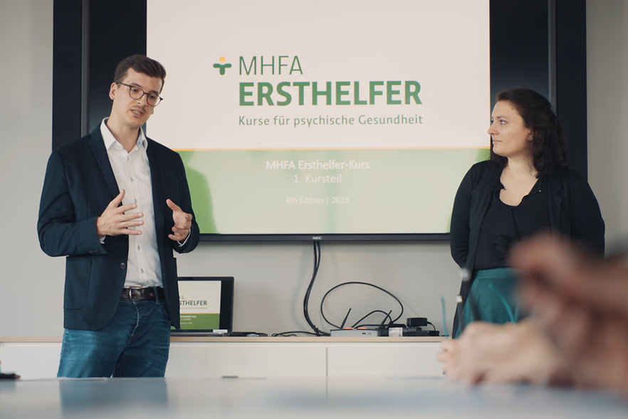 Zwei Menschen stehen vor einer Leinwand mit der Aufschrift "MHFA Ersthelfer" 