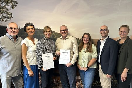 Eine Gruppe von Personen steht aufgereiht vor einer Wand und freut sich &uuml;ber zwei Urkunden zur Schulpatenschaft.