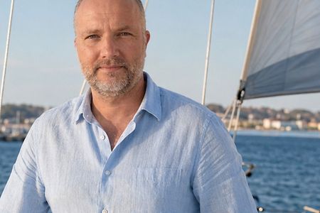 Lars von Osten auf einem Segelboot bei sonnigem Wetter, im Hintergrund Wasser, weiter weg sieht man Hafen und H&auml;user.