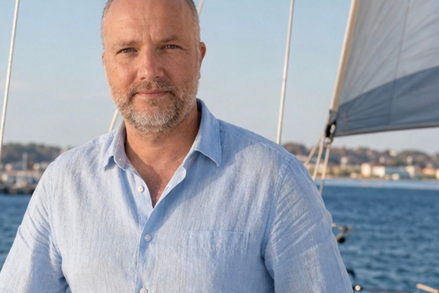 Lars von Osten auf einem Segelboot bei sonnigem Wetter, im Hintergrund Wasser, weiter weg sieht man Hafen und H&auml;user.