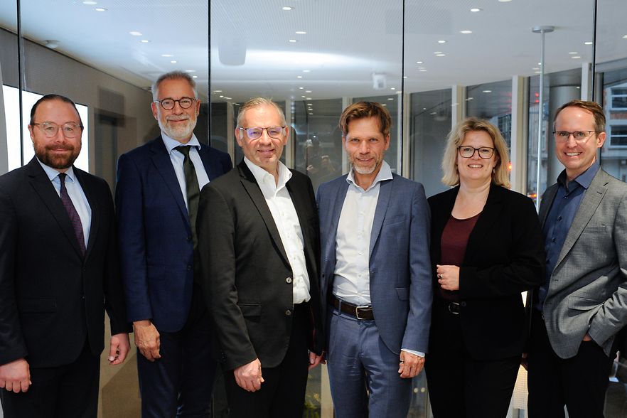 F&uuml;nf M&auml;nner und eine Frau stehen vor einem Konferenzraum und l&auml;cheln in die Kamera.