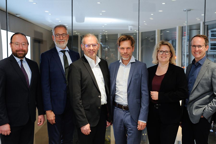 Fünf Männer und eine Frau stehen vor einem Konferenzraum und lächeln in die Kamera.