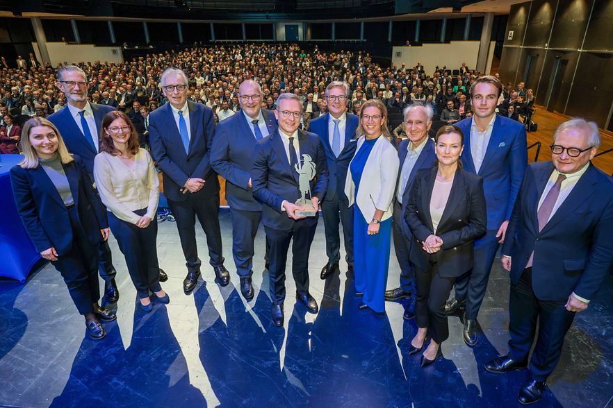 Zw&ouml;lf Personen posieren mit einer Auszeichnung vor einem voll besetzten Saal.