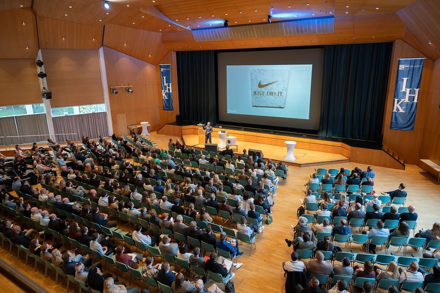 Das Bild zeigt den gro&szlig;en Veranstaltungssaal aus der Vogelperspektive. Vorne steht ein Redner vor einer PowerPoint.