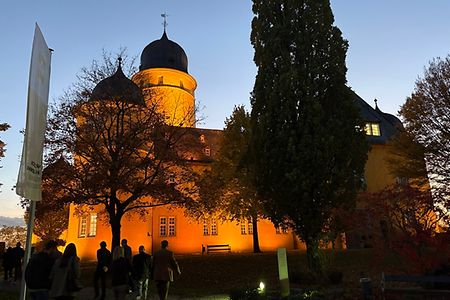 Ein beleuchtetes gelbes Schloss in der Abendd&auml;mmerung, davor eine laufende Personengruppe.